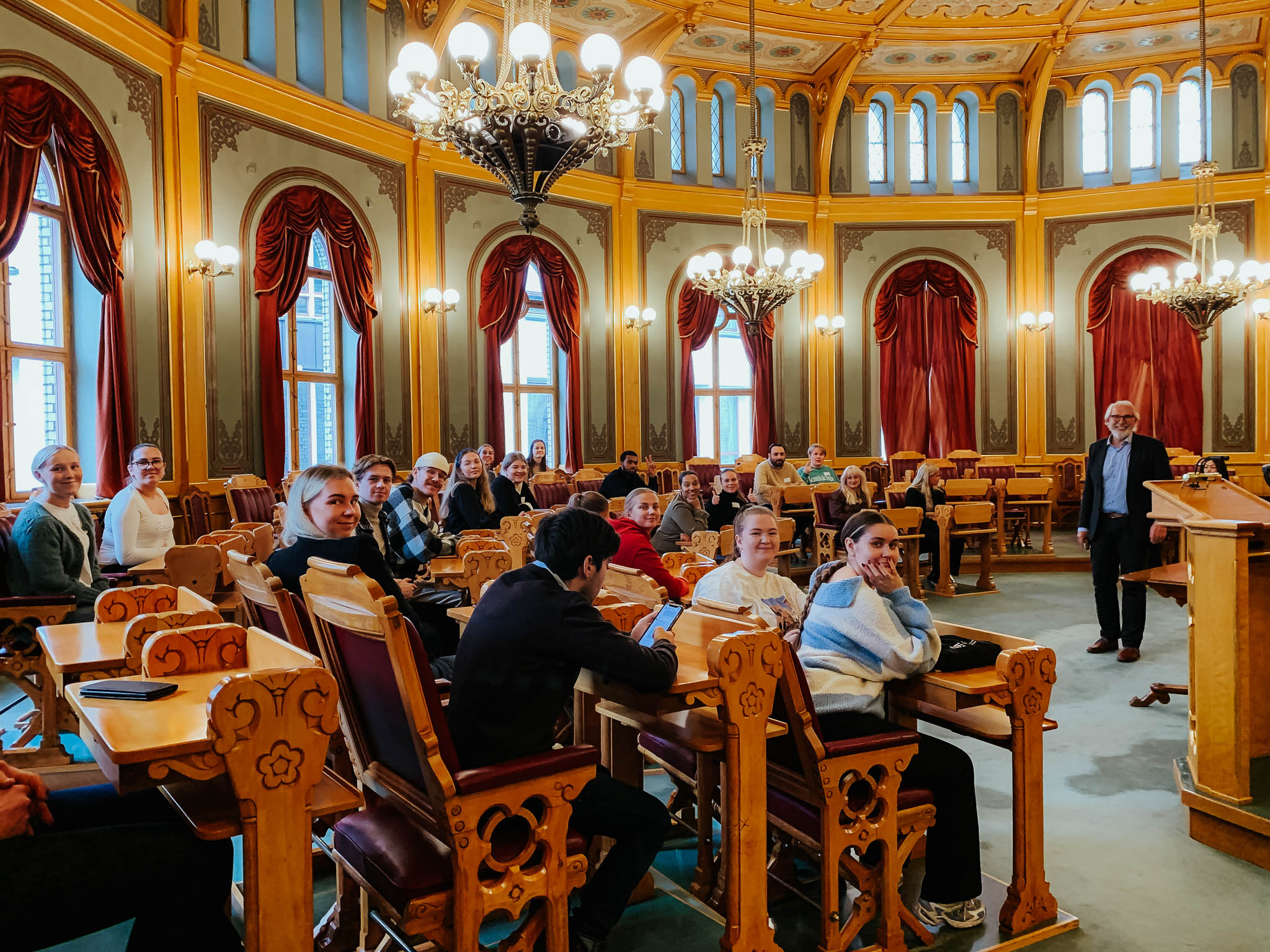 En gruppe studenter er plassert i stortingssalen sammen med Carl Erik Grimstad. De smiler mot kamera. Foto.