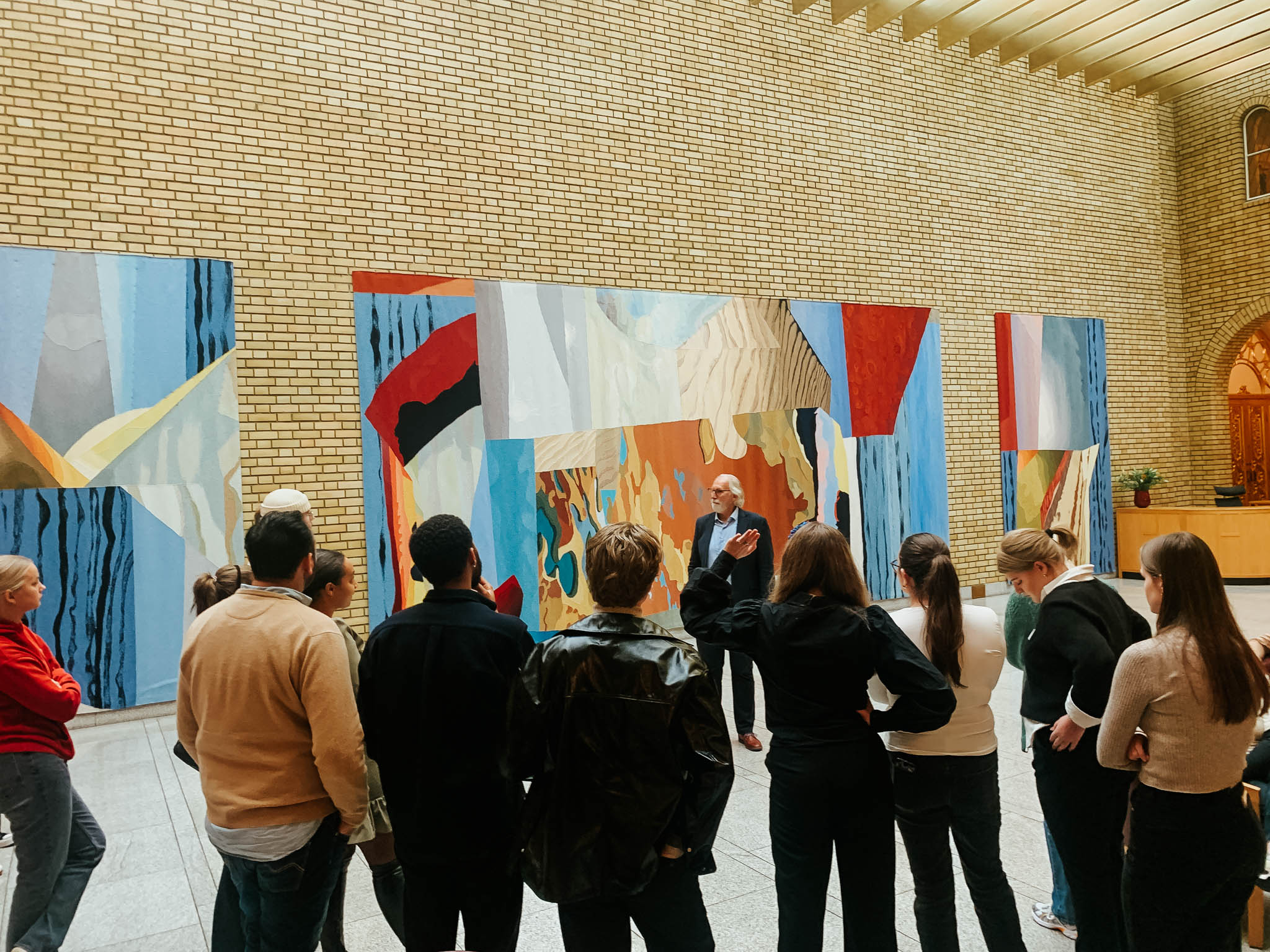 En gruppe studenter står inne på Stortinget med ryggen vendt mot kamera. De ser mot Carl Erik Grimstad som forteller om kunsten på veggene. Foto.