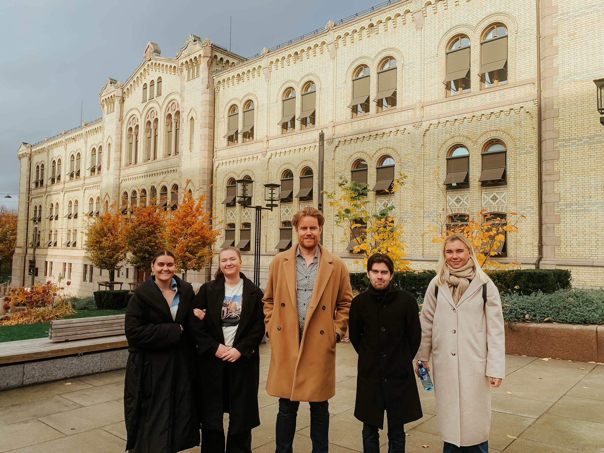 Fem studenter står foran langsiden av Stortinget mens de smiler mot kamera. De har på seg høstklær. Foto.