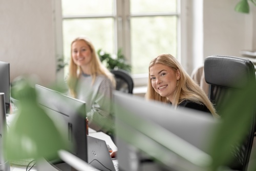 To personer sitter i et lyst kontormiljø på PC og smiler i kamera