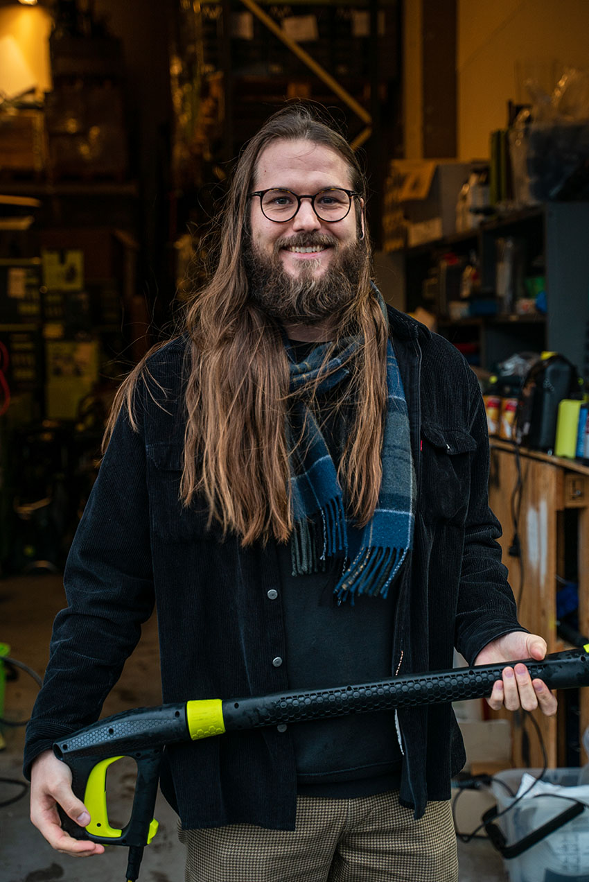 Christopher Harestad står utenfor lagerporten til AVA of Norway. Holder en høytrykkspyler i hendene. Ser i kamera og smiler.