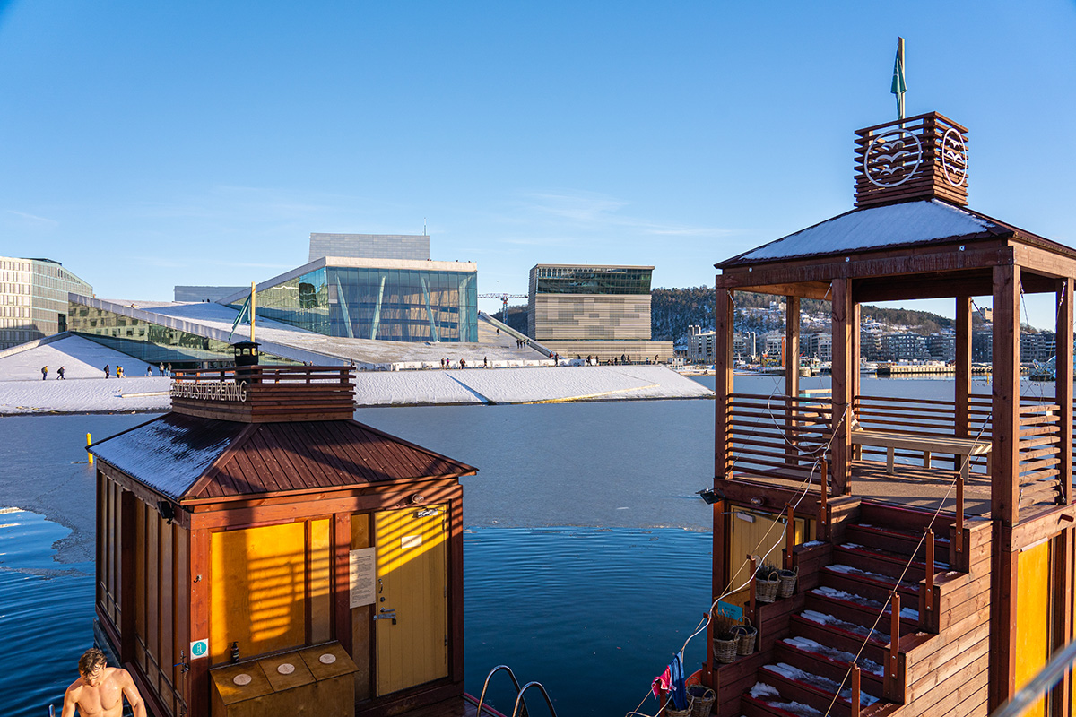 Fargerike flytende badstuehytter på vannet med person på dekk, med utsikt over det moderne Operahuset i Oslo og byens silhuett.