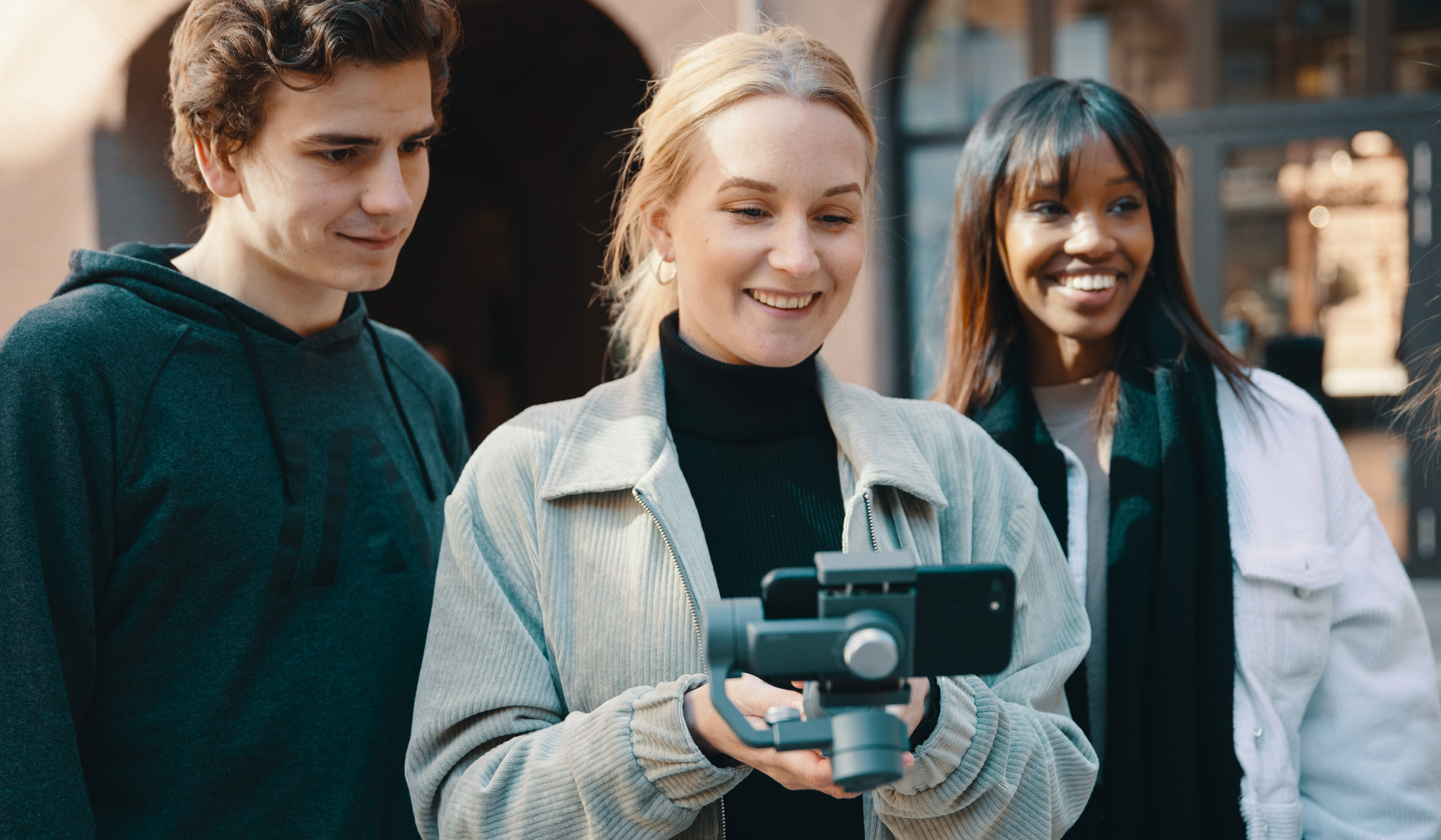 Student på Digital innholdsproduksjon, Maja Granli Jensen lærer seg å bruke mobil til å lage videoer. Hun har blondt hår og beige jakke, og filmer med mobiltelefon. Hun står med to klassekamerater, en gutt og en jente. De smiler og ser på mobiltelefonen mens de filmer. 