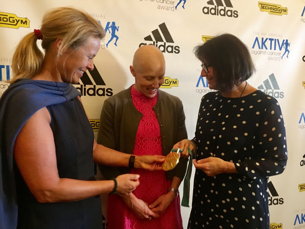 Kreftrammet kvinne viser fram en gullmedalje til to andre kvinner.