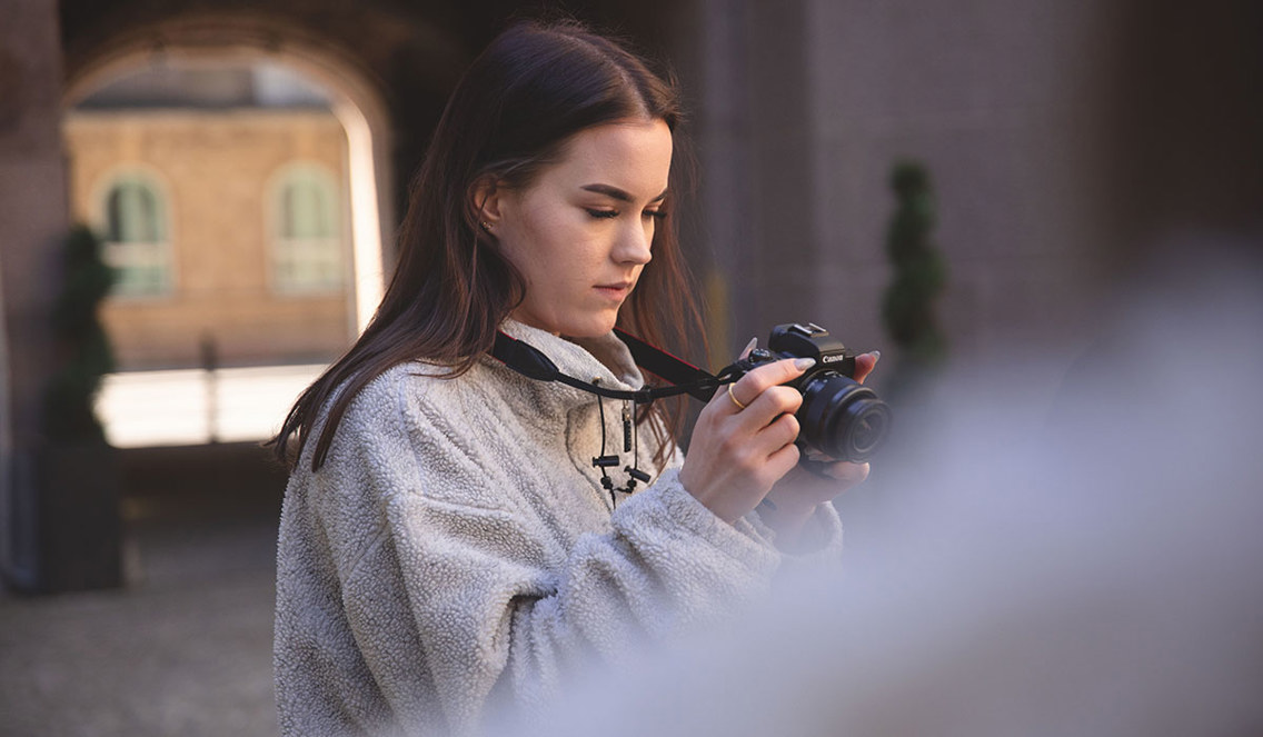 En kvinnelig student på Fagskolen Kristiania studerer kameraet sitt. Hun har langt mørkt hår og en grå fleece-genser.