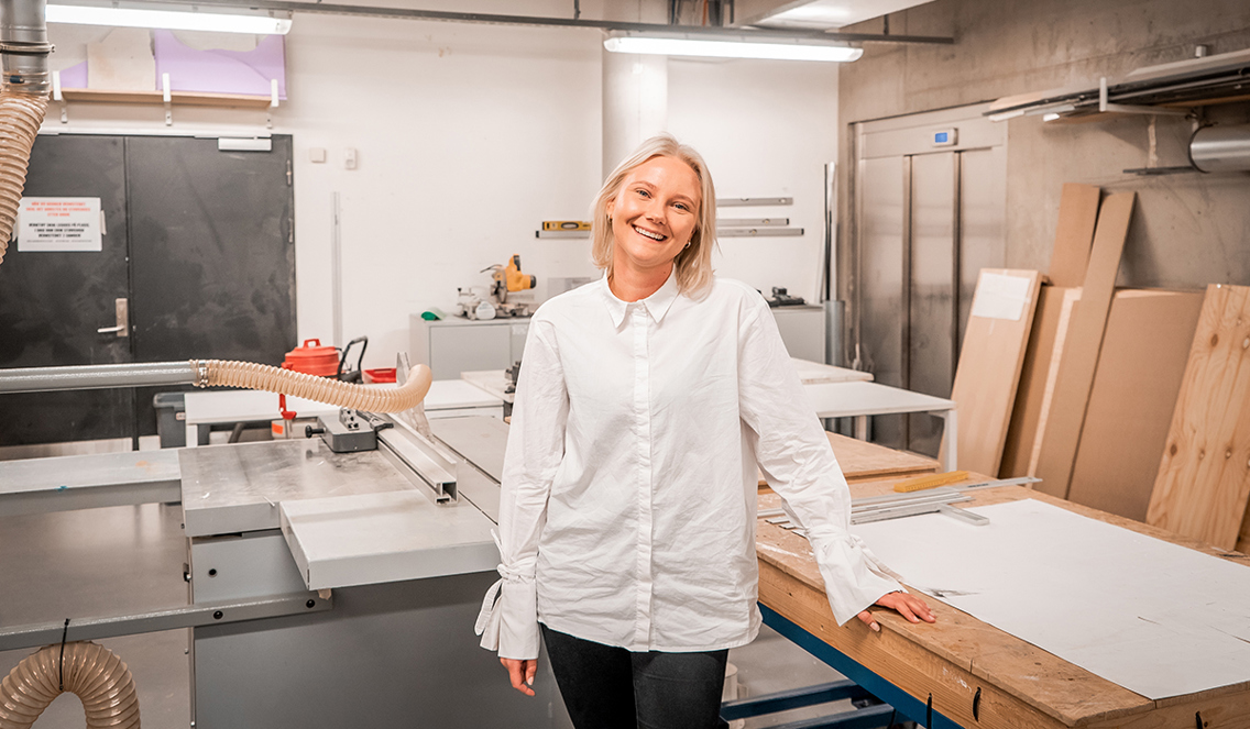 Ida står i et makersspace på campus Vulkan. Hun har på seg en hvit bluse og holder den ene hånden på en arbeidsbenk bak henne. 