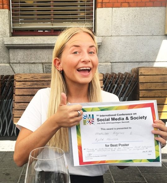 Mathilde her med diplomet for Best Poster. Diplomet er fargerikt og har hint av rødt, grønt og gult. Mathilde har langt lyst hår og holder tommelen opp. Hun ser strålende fornøyd ut.