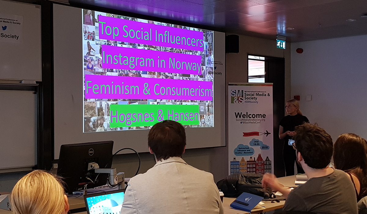 Mathilde Hogsnes presenterer under International Conference on Social Media and Society Awards i København. Hun står foran et lærret som har oppgaven hennes på skjermen. Skjermen er i rosa neon-skrift. 