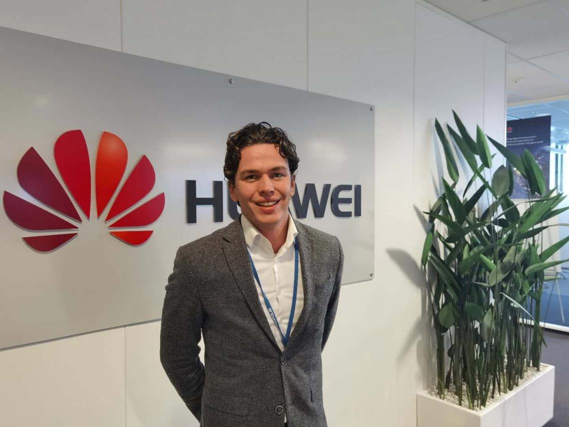 Cedric står foran en Huawei-logo i deres lokaler. Han er iført dress og nøkkelkort rundt halsen. 