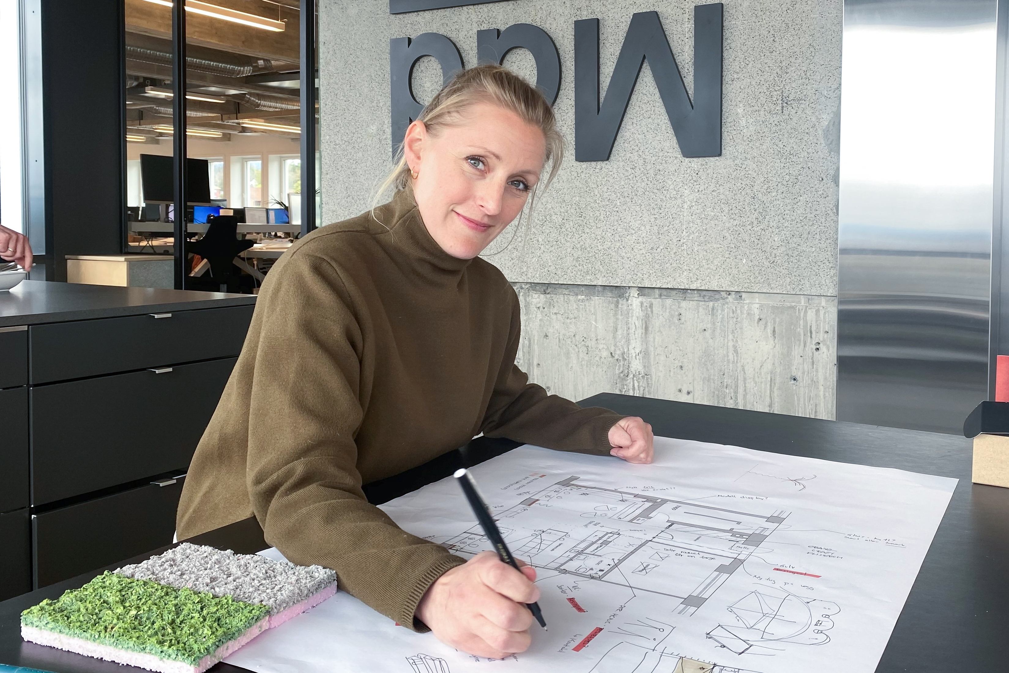 Synne sitter ved en pult på kontoret, og tegner på en plantegning.
