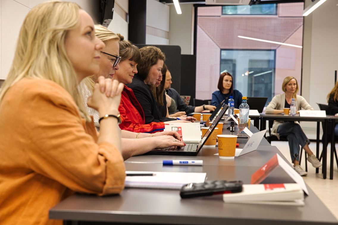 Voksne studenter sitter i klasserom med pultene i hestesko og lytter til foreleser.
