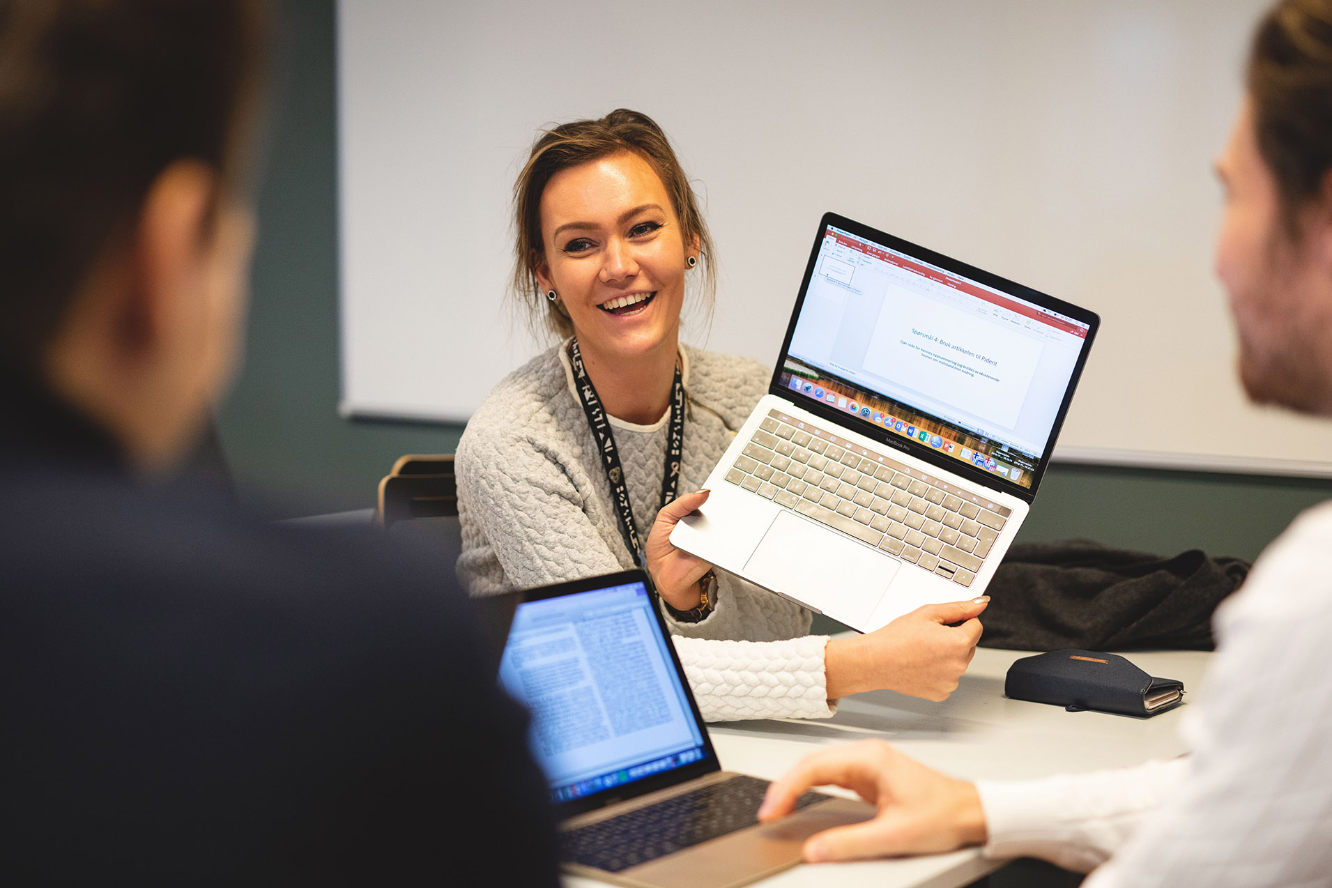 Foto av kvinnelig student som holder opp en laptop for å vise skjermen til to mannlige medstudenter.