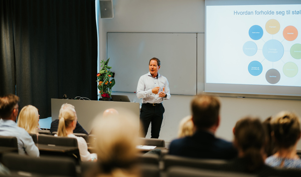 Kalhovde står foran i et auditorium og snakker til mange personer, som sitter med ryggen til. 