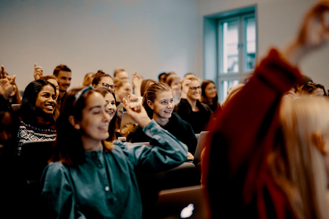 Studenter i et klasserom. Noen smiler og noen rekker opp hånden