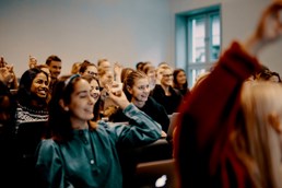 Studenter i et klasserom. Noen smiler og noen rekker opp hånden