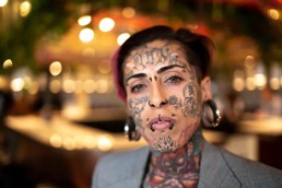 Kvinne med kort hår og dress med flere tattoveringer og piercinger i ansiktet ser rett inn i kamera