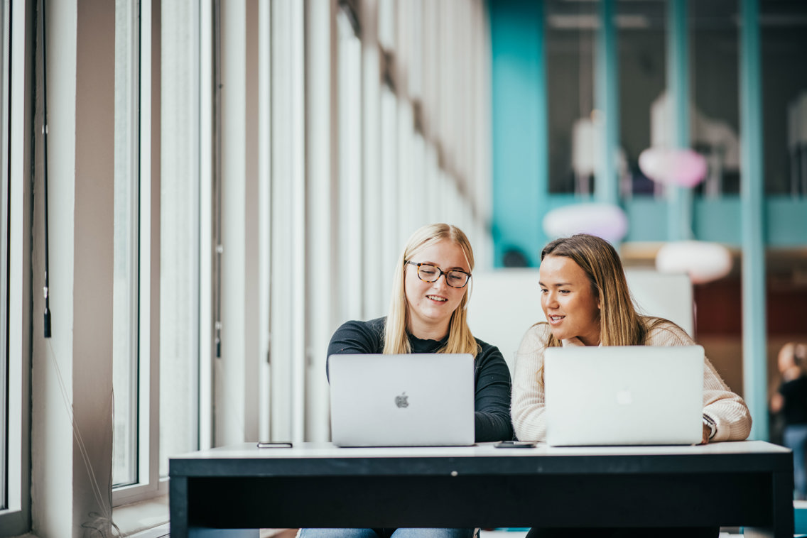 To kvinnelige studenter sitter i et lyst lokale med hver sin mac.