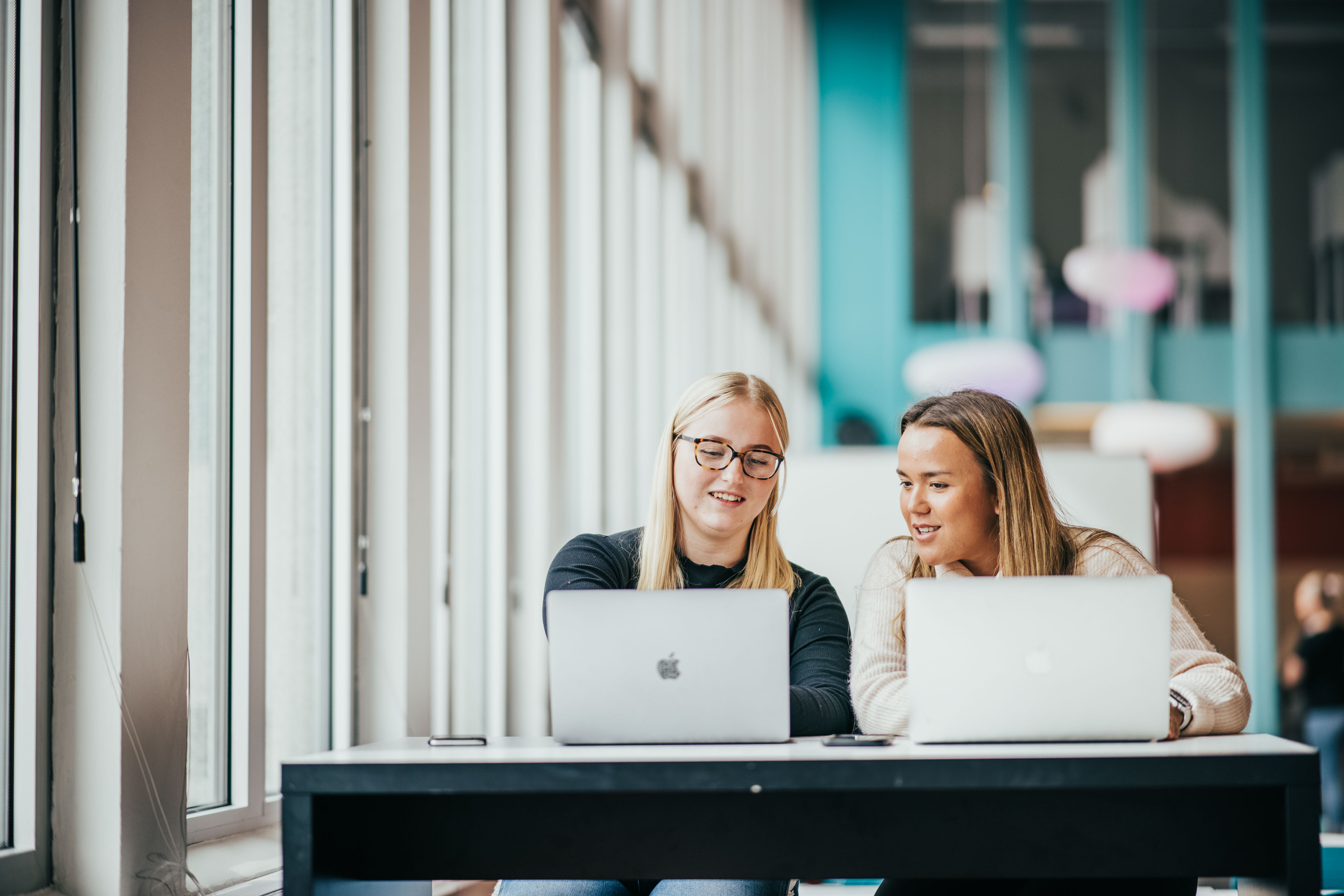 To kvinnelige studenter sitter i et lyst lokale med hver sin mac.