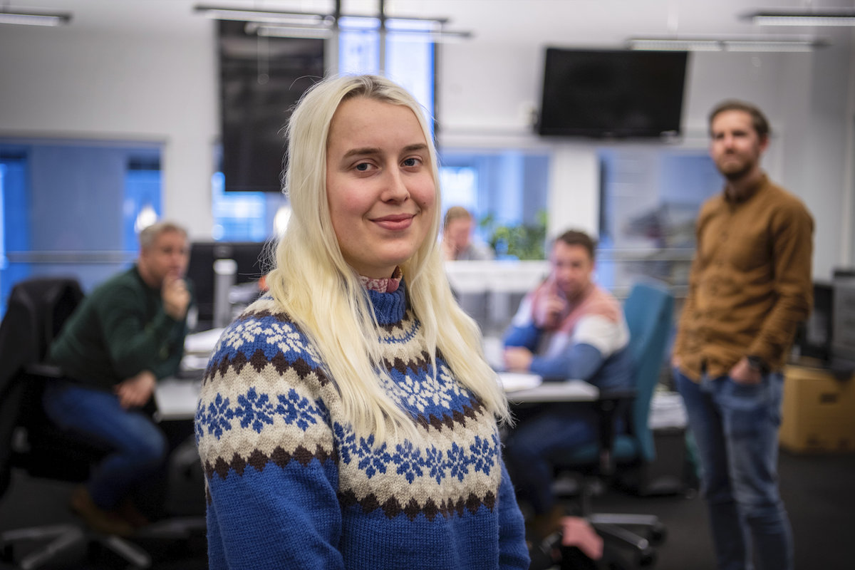 Maria står i strikkegenser i redaksjonen i Hamar Arbeiderblad. Bak henne står det noen kolleger. 