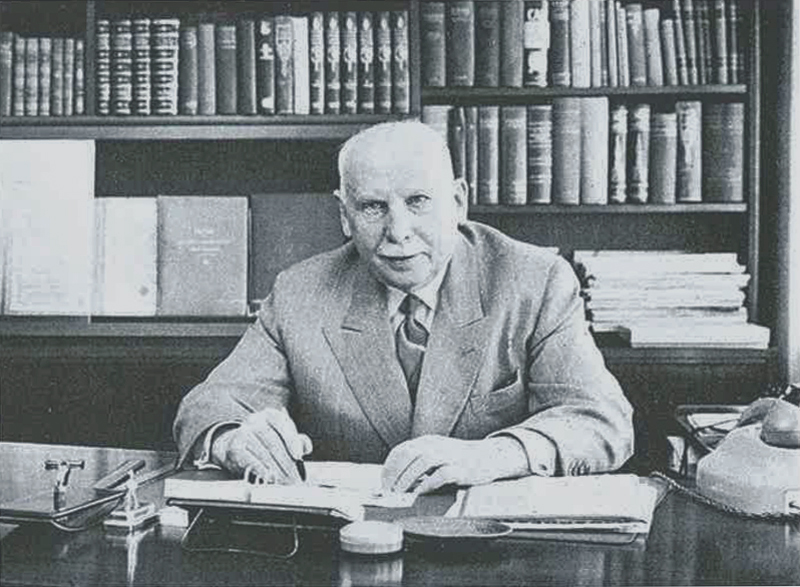 Foto i sort hvitt av en mann i dress som sitter ved pult i et arbeidsrom.