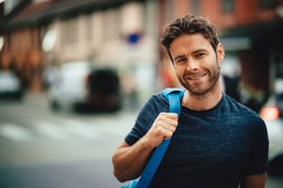 En mann med mørkt hår smiler til kamera. Han står utendørs og har en blå ransel over den ene skulderen