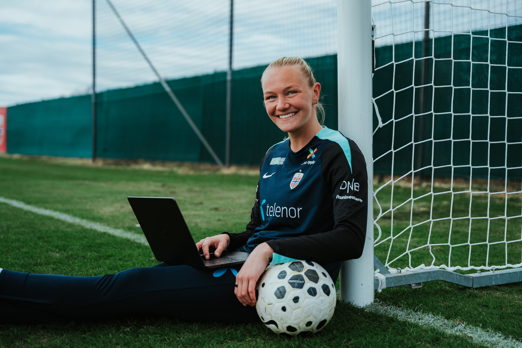 Frida sitter på banen ved målet og studerer på Macen. Ved siden av henne ligger en fotball.