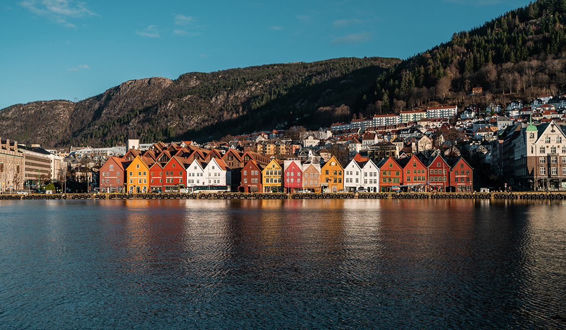 Bildet viser et bilde av Bryggen i Bergen. Solen stråler på de gamle husene og himmelen er blå. 