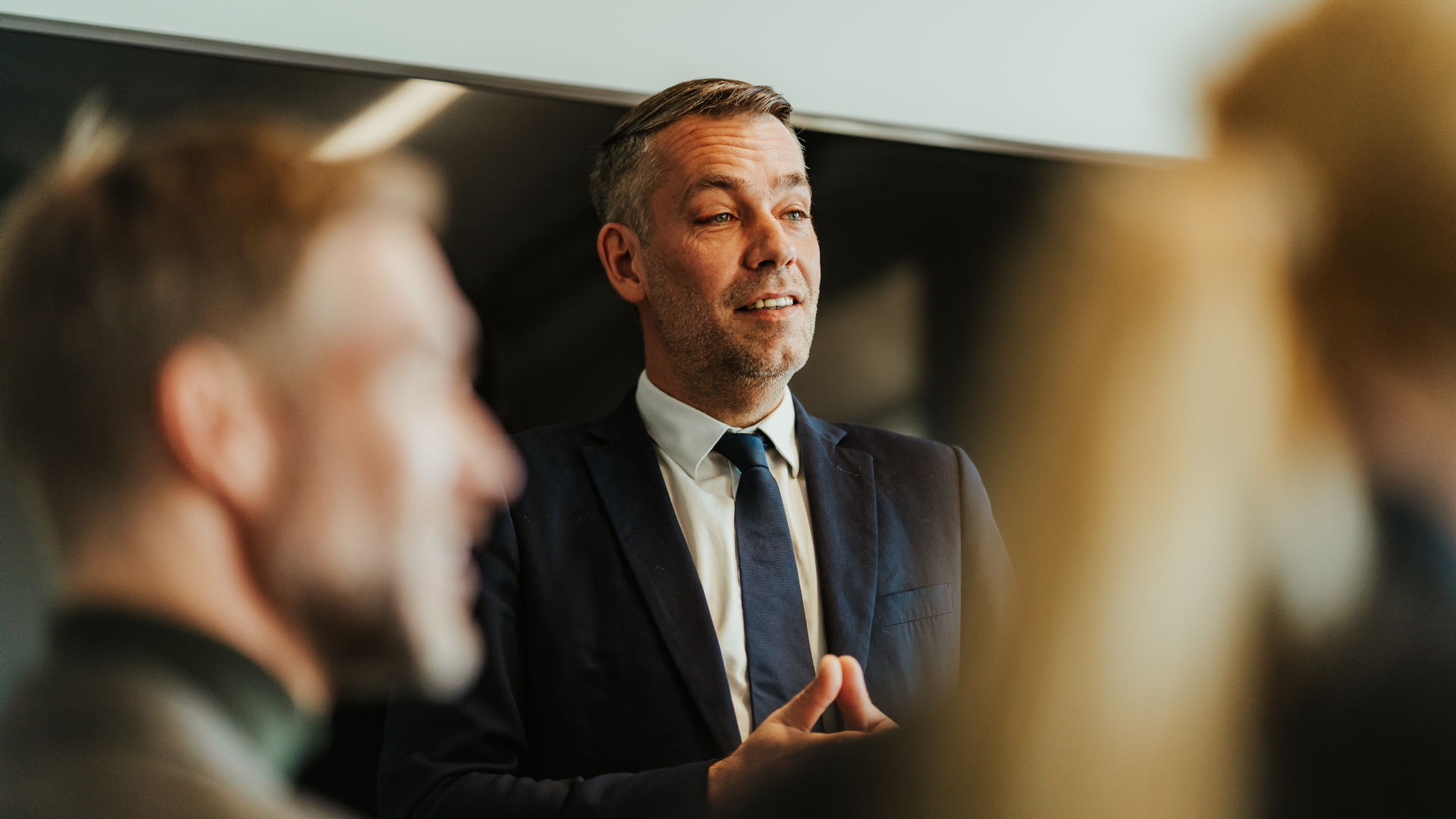 Mann står i et møterom og snakker i et møte. To personer sitter med ryggen til.