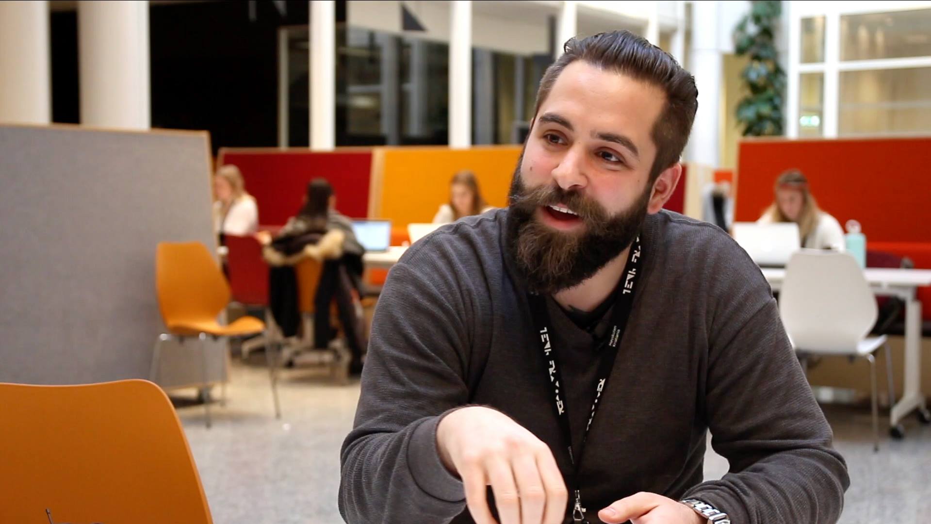 Mann med skjedd og grå genser sitter ved et bord og prater engasjert