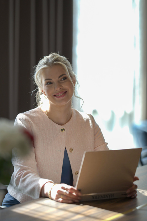 Celine sitter ved bordet foran PC-en og smiler, med et vindu og gardiner i bakgrunnen.