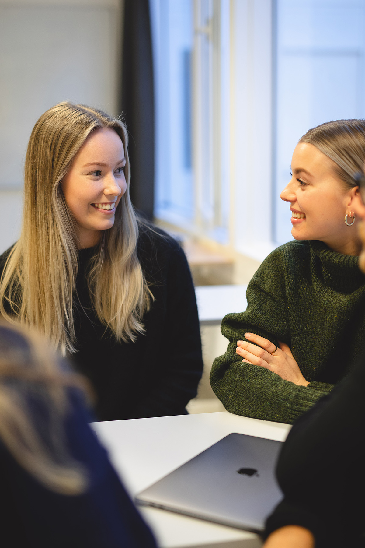 To kvinnelige studenter sitter foran et bord og smiler mot hverandre. Den ene har langt lysebrunt hår og den andre har brunt hår oppsatt i en dott. 