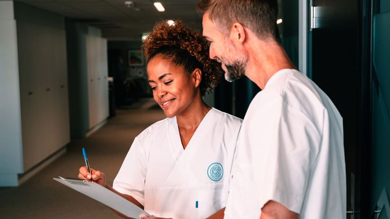 To helsearbeidere i hvite uniformer står i en korridor og ser på en dokumentmappe. Den kvinnelige helsearbeideren holder en penn og noterer noe, mens den mannlige helsearbeideren smiler