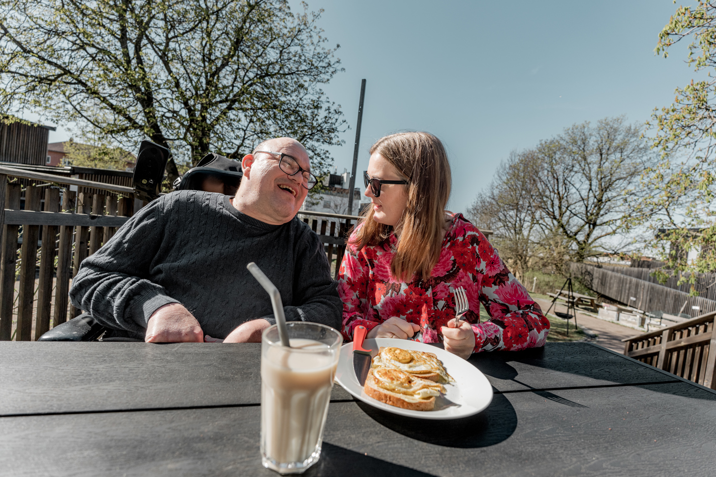 To personer sitter ved et utebord og spiser i solfylt omgivelser, og smiler i samtalen. 