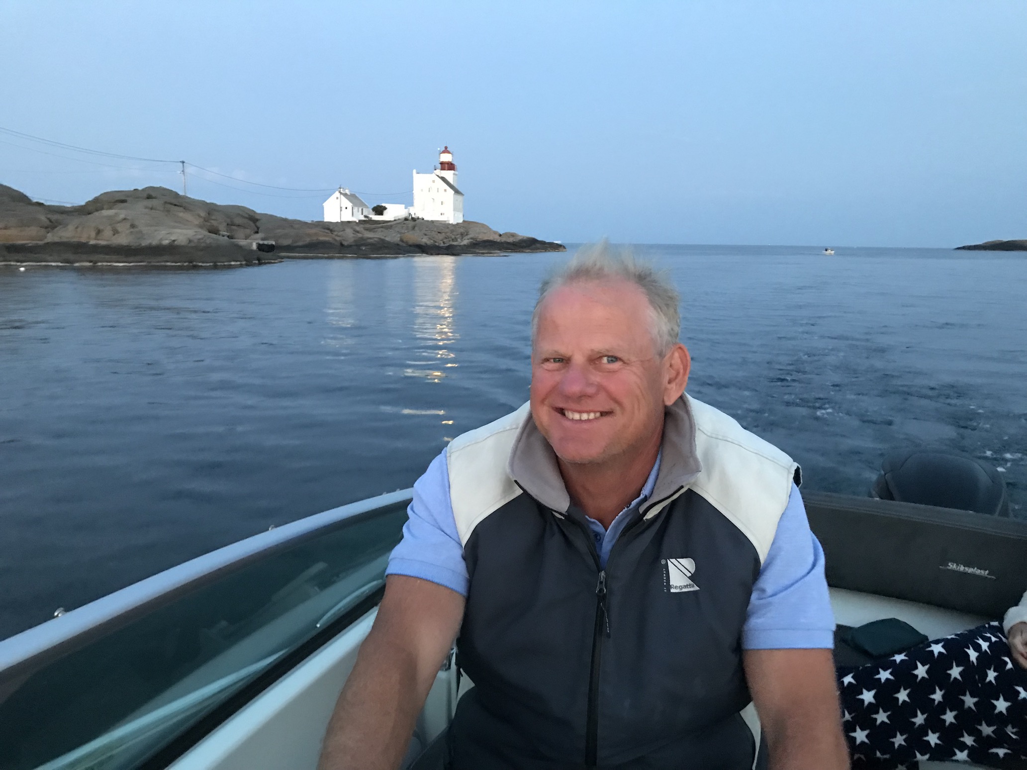Nils Høgevols sitter i motorbåt med et stort smil. Rundt han er sjøen og i bakgrunnen er en odde med et fyr