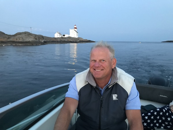 Nils Høgevols sitter i motorbåt med et stort smil. Rundt han er sjøen og i bakgrunnen er en odde med et fyr