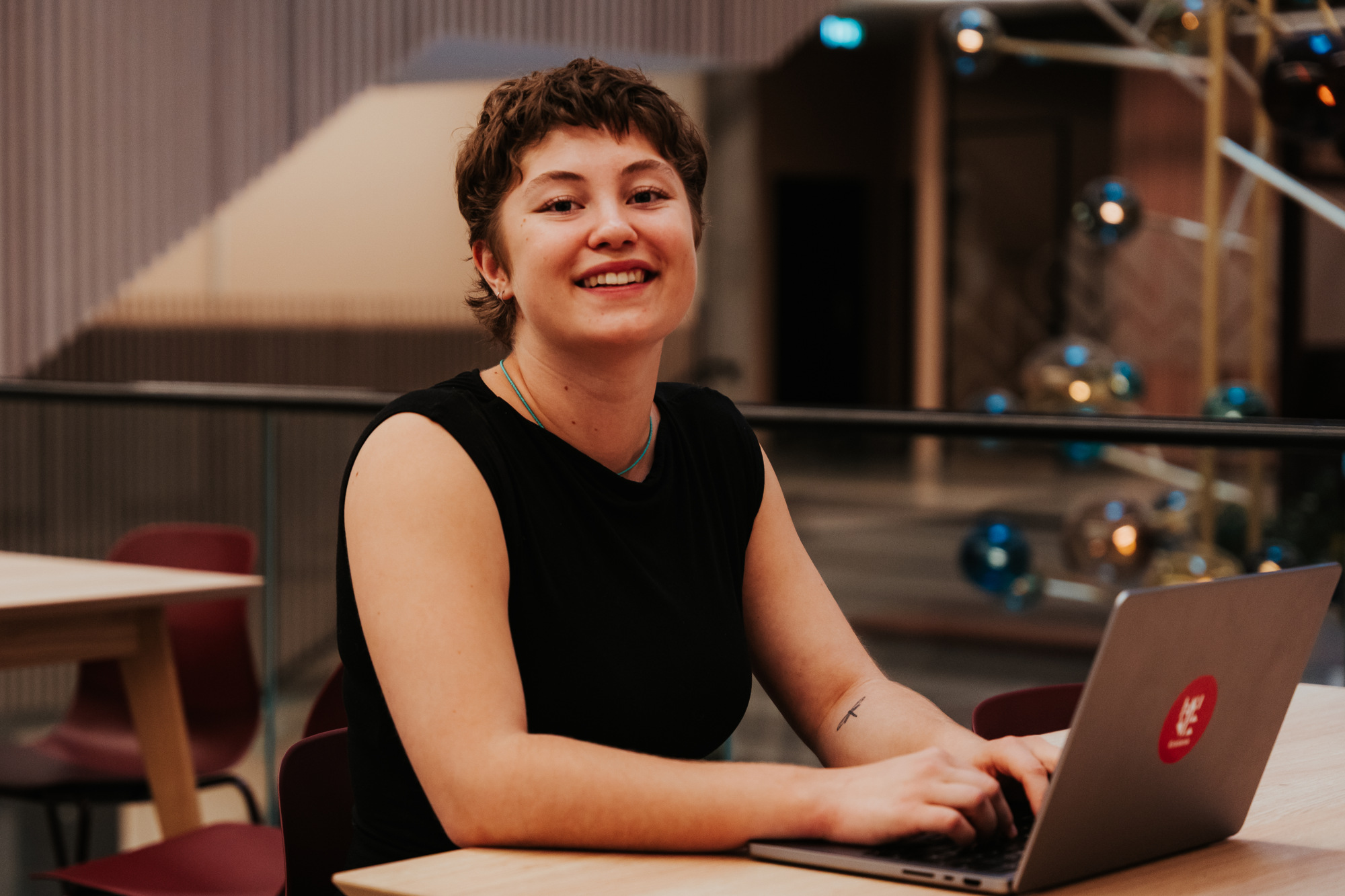 En student sitter ved en laptop og smiler i et moderne læringsmiljø.