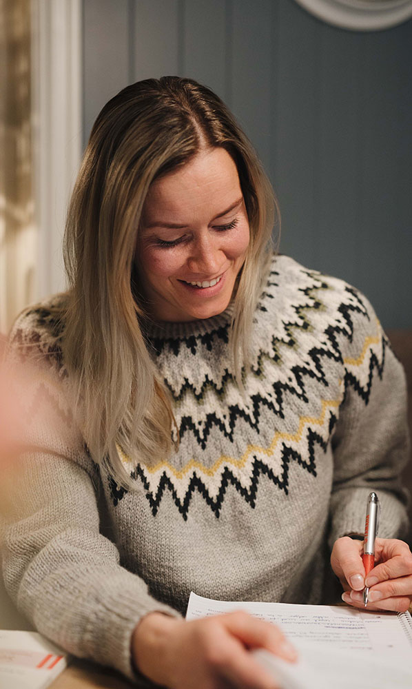 Ågot iveren sitter på knøkkenet hjemme og gjør lekser til nettstudier på Fagskolen Kristiania. Hun har på seg en  mønstrete grå ullgenser. Hun smiler men hun skriver notater .