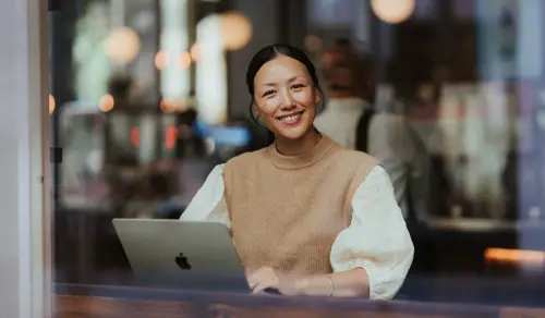 Kvinne sitter og studerer på en mac, hun smiler til kamera.