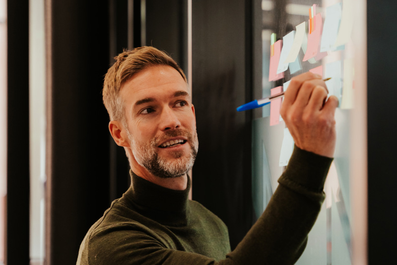 Foto av mann i høyhalset genser som skriver på post it-lapp på glassvegg.
