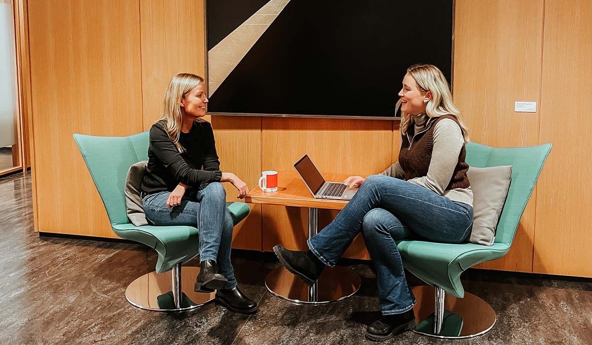 Selma Vedum Håland og Helle Campbell  sitter i hver sin turkise stol mens de smiler til hverandre. På det runde bordet mellom de står det en bærbar PC. Foto.