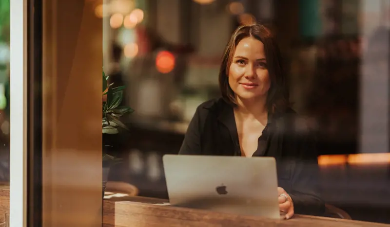 Kvinne sitter på cafe og jobber på en mac. Hun smiler til kamera.