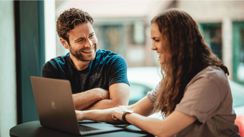 To studenter smiler og samarbeider rundt en laptop ved et bord, i et lyst rom med store vinduer