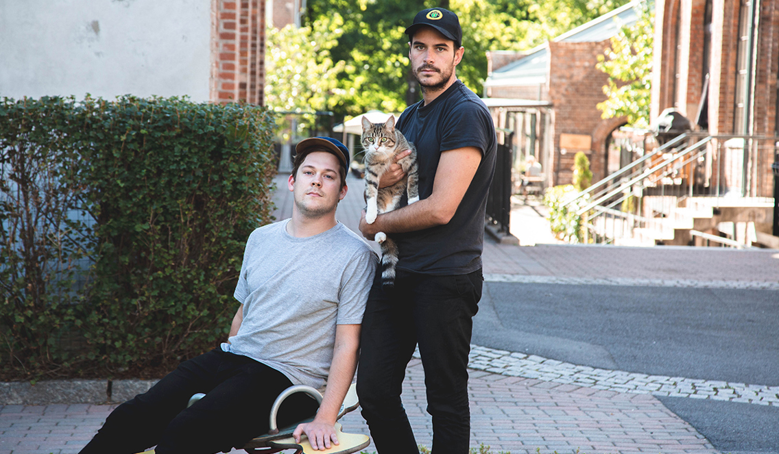 To unge menn står på en gate. Den ene gutten holder en katt. Den andre gutten sitter på en gyngeleke. 