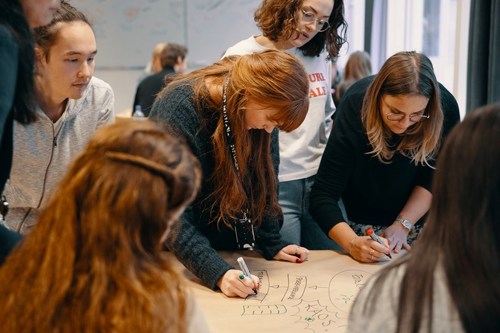 Foto av en gruppe studenter som står samlet rundt et bord og skriver på et gråpapir.