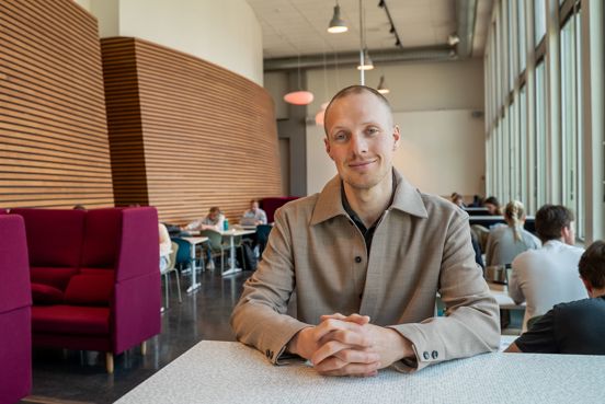 Robin Bjorheim sitter bak et bord i et atrium, med studenter i samtale bak seg.