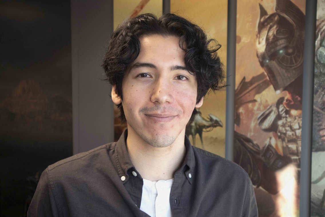 Nærbilde av en smilende Henry Ramsevik i lokalene til Funcom. I bakgrunner ser man et bilde fra et av spillene til Funcom, det viser en mann med hjelm og rustning. 