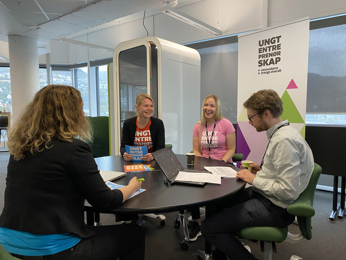 Fire representanter fra Ungt Entreprenørskap sitter rundt et bord med logoen bak seg.