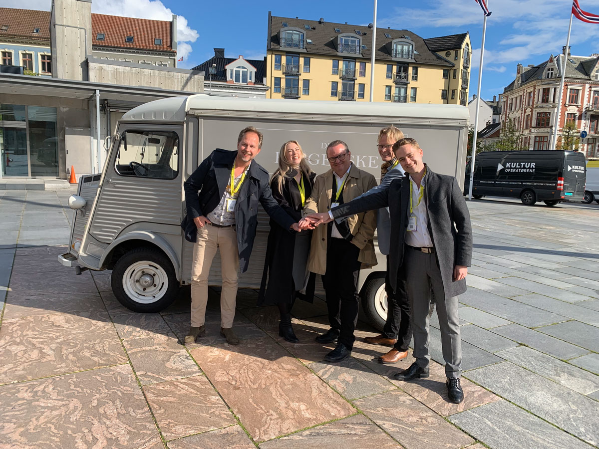 Fem personer står samlet på en åpen plass i solskinn. De står som et team i en halvsirkel med hendene lagt oppå hverandre.