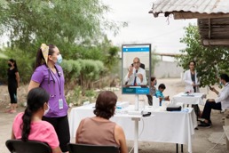 Woman with a mask standing next to a screen outside with two patients sitting opposite listening to the doctor on the screen