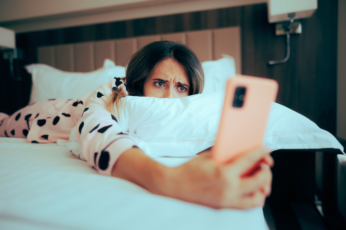 Woman laying in her bed looking worringly on her phone.